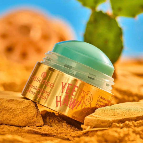 Un Essence Primer en Stick Hydratant Base Longue Tenue 10,2 g, dont l'emballage or et turquoise porte la mention "YEE HAW !", repose sur du sable doré, dans un décor de verdure floue et de ciel bleu vif. Dakar