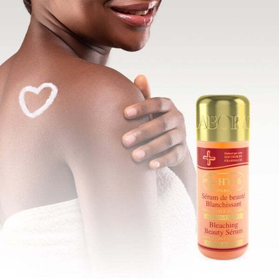 Une femme souriante à la peau foncée et au cœur blanc sur l'épaule se tient près d'un flacon de "Ht26 Sérum de Beauté Blanchissant Corps Huile de Carotte 150 ml", enveloppé dans une serviette blanche sur un fond clair. Dakar