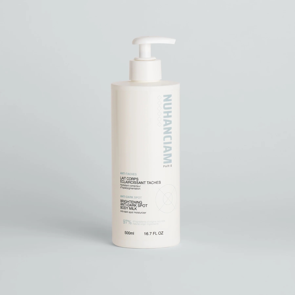 Un grand flacon pompe blanc étiqueté "Nuhanciam Paris" contient le Lait Corps Eclaircissant Shot d'Univers Cosmetix, conçu pour lutter contre les taches brunes. Le flacon de 500 ml se détache sur un fond clair uni. Dakar