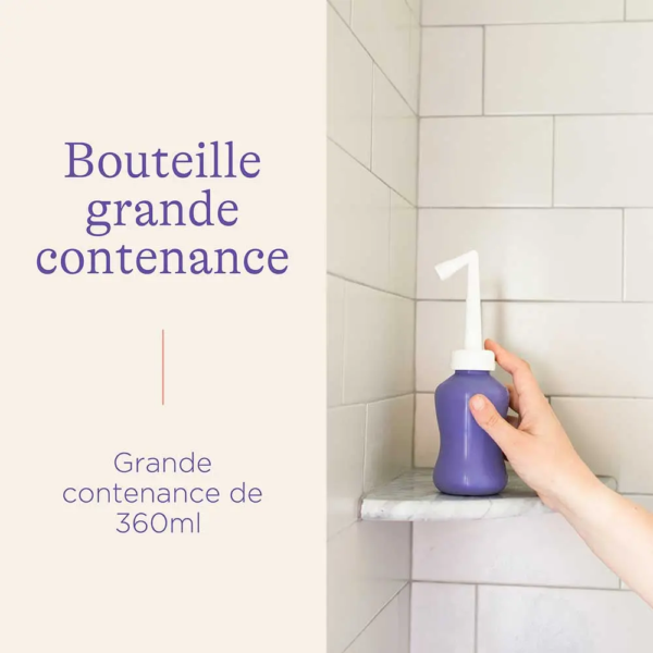 Une main tient une pièce de Lansinoh Douche Intime Périnée 1 - un flacon violet avec un embout angulaire blanc - sur une étagère de douche. Le texte indique : "Bouteille grande contenance" et "Grande contenance de 360ml. Dakar