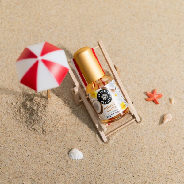Un flacon de 30 ml de l'Eau de Toilette Les Petits Plaisirs Monoï Coco est posé sur une mini chaise de plage dans le sable, à côté d'un petit parasol rouge et blanc, avec des coquillages et des étoiles de mer à proximité. Dakar