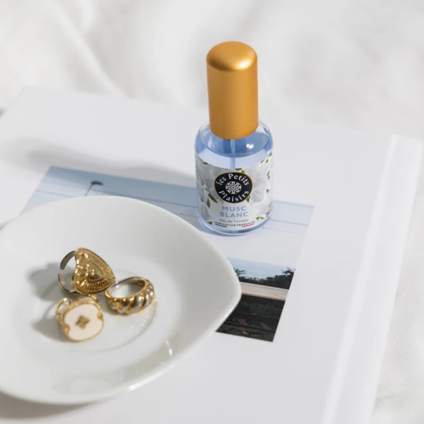 Une petite coupelle blanche avec trois anneaux en or côtoie un flacon de Les Petits Plaisirs Eau de Toilette Musc Blanc 30 ml, le tout posé sur un livre blanc dont la couverture est ornée d'une photo de paysage. Dakar
