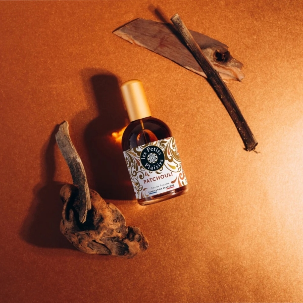 Un flacon de 110 ml de l'Eau de Toilette Patchouli Les Petits Plaisirs avec un bouchon doré repose sur une surface en cuivre, entourée de bois et d'écorces pour un aspect chaud et terreux. Dakar