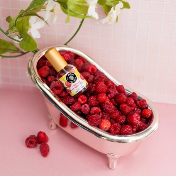 Une petite baignoire métallique remplie de framboises fraîches, de fraises et de Les Petits Plaisirs Eau de toilette Vanille Framboise 30 ml est posée sur une surface rose avec des fleurs et un mur carrelé rose en arrière-plan. Dakar