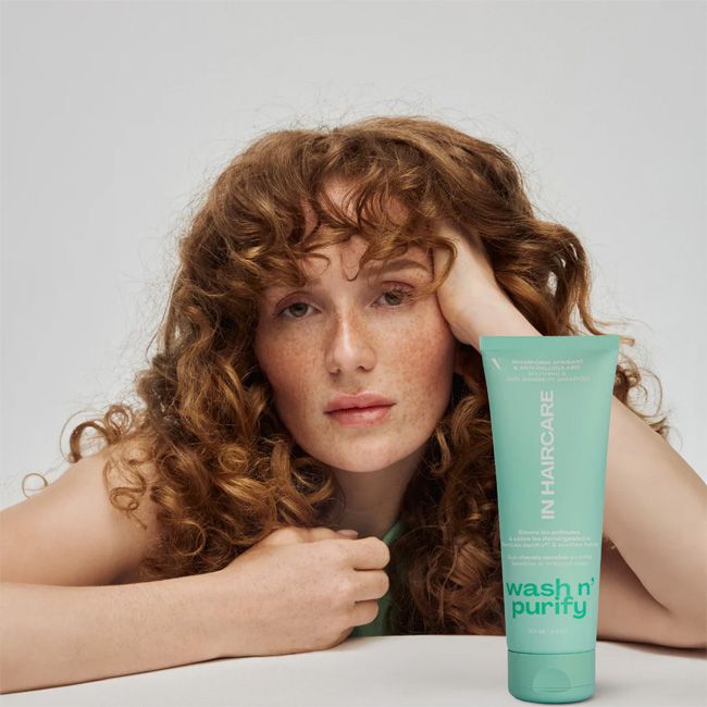 Une femme aux cheveux roux bouclés et aux taches de rousseur s'appuie sur une surface blanche, en regardant la caméra. Devant elle, un tube sarcelle de In HairCare Shampoing Apaisant Wash n' Purify Purifie et Rééquilibre 250ml sur un fond gris clair. Dakar