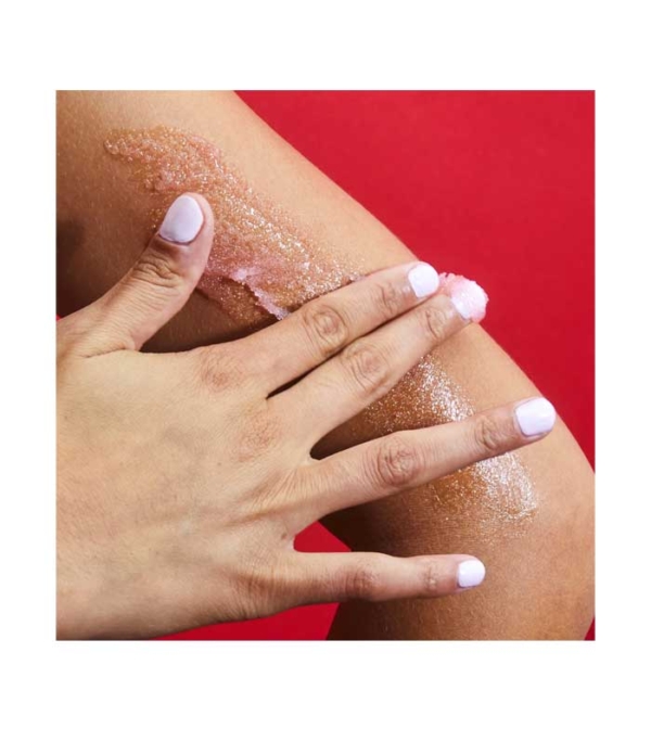 Une main aux ongles rose pâle applique Gommage corporel à la fraise et au sucre de karité 510 g, un exfoliant granuleux, sur le genou d'une personne, sur un fond rouge vif. Dakar