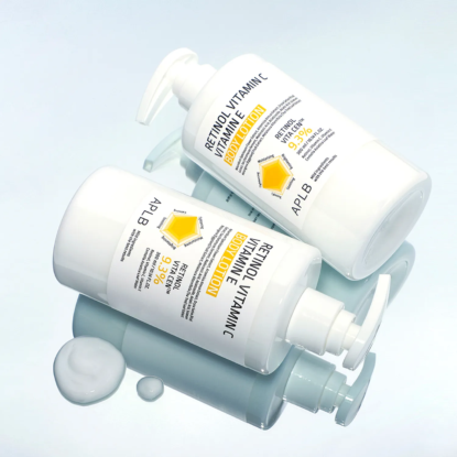 Deux flacons à pompe blancs étiquetés "Retinol Vitamin C Hydration" de Univers Cosmetix avec des accents jaunes reposent sur une surface bleue claire et brillante. De la lotion blanche est renversée à proximité. Les deux flacons portent le logo de la marque APLB. Dakar