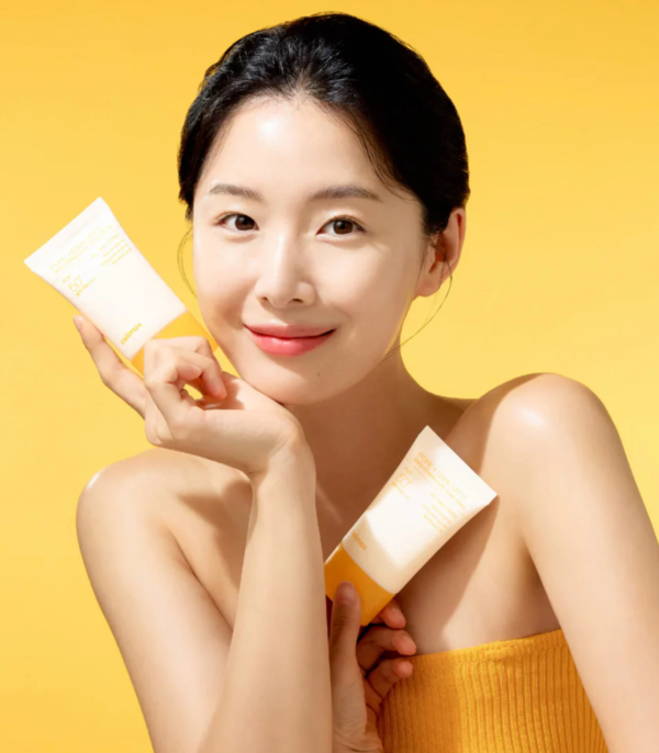 Une femme à la peau claire et aux cheveux foncés sourit, tenant deux tubes de produit de soin sur un fond jaune vif. Elle porte une serviette jaune et semble avoir le visage frais, ce qui lui confère un aspect propre et dynamique. Dakar
