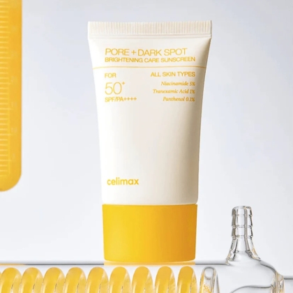 Un tube de crème solaire celimax Pore + Dark Spot Brightening Care avec SPF 50+ est présenté à la verticale sur un fond clair, avec un emballage jaune et blanc. Des objets en verre et un élément jaune sont partiellement visibles à proximité. Dakar