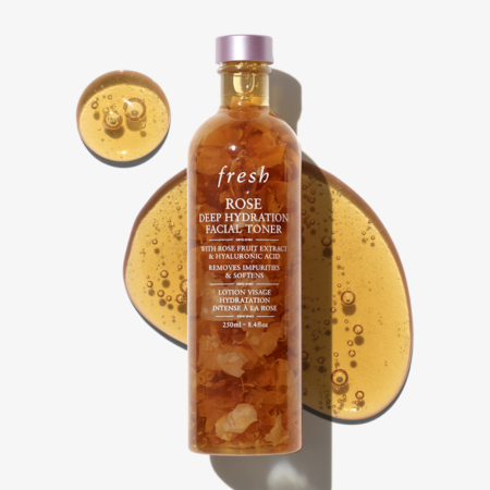 Un flacon transparent de Fresh Rose Deep Hydration Lotion hydratation Intense à la Rose 250 ml avec des pétales de rose visibles repose sur une surface blanche, entouré de deux flaques de liquide doré et de son ombre. Dakar