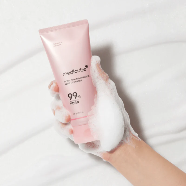 Une main couverte de mousse blanche tient un tube rose de Medicube Pore Pink Niacinamide Whip Cleanser, avec 99% pore care écrit sur le tube, sur un fond blanc et mousseux. Dakar