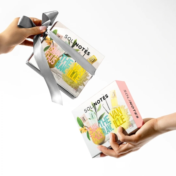Deux mains s'échangent un coffret Solinotes Découverte Osmanthus Thé Blanc Vanille, décoré d'un ruban argenté et d'images vibrantes des trois parfums sur un fond blanc éclatant. Dakar