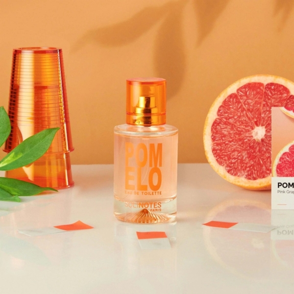 Un flacon de Solinotes Eau De Parfum Pomelo 50 ml pour Femme repose sur une surface blanche avec des coupes orange, des feuilles vertes, un pamplemousse rose coupé en deux et sa boîte sur un fond pêche. Dakar