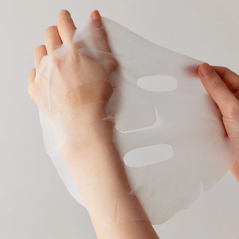 Une main tient un masque facial translucide avec des découpes pour les yeux, le nez et la bouche sur un fond neutre. Dakar