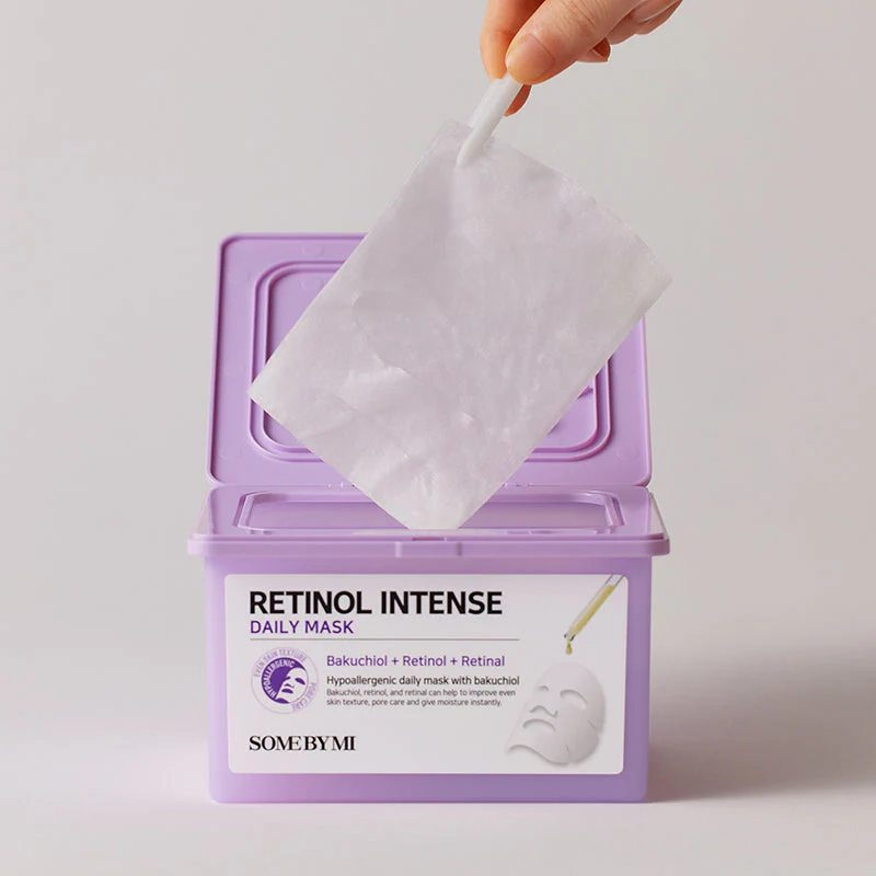 Une main tire un masque en feuille d'un récipient lavande étiqueté "Retinol Intense Daily Mask" par SOME BY MI, avec un diagramme d'un masque et des détails sur les ingrédients sur le devant. Dakar