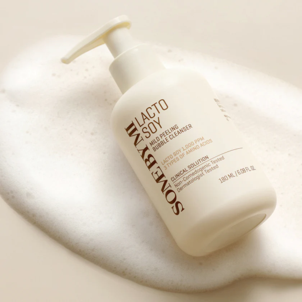 Un flacon pompe blanc de Some By Mi Nettoyant Moussant et Exfoliant Doux Lacto Soy 180ml repose sur une surface mousseuse et texturée. L'étiquette affiche un texte en or et en marron. Dakar