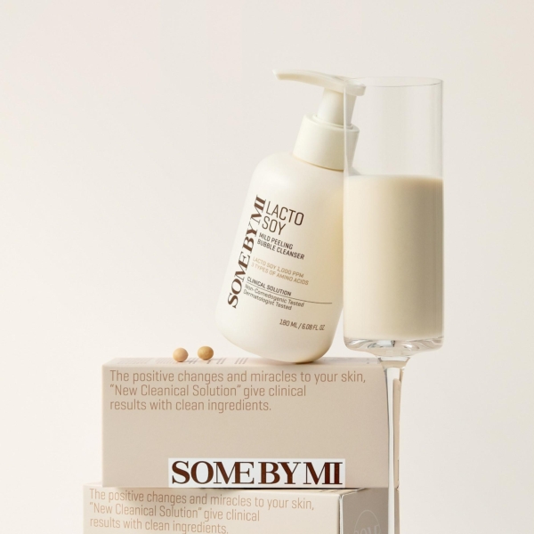 Un flacon de Some By Mi Nettoyant Moussant et Exfoliant Doux Lacto Soy 180ml est posé sur une boîte de marque à côté d'un grand verre de liquide laiteux, avec deux perles beiges sur la boîte sur un fond clair et minimaliste. Dakar
