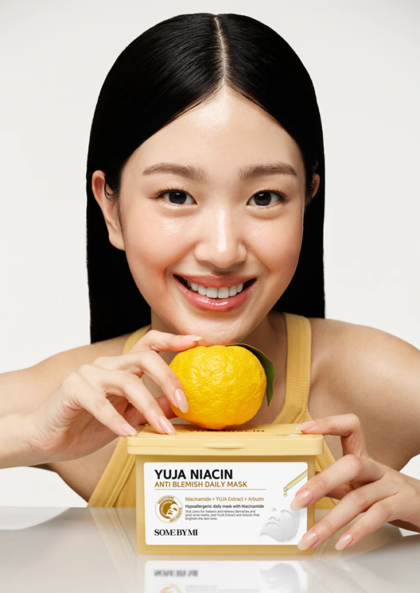 Une femme souriante vêtue d'un haut jaune tient un agrume jaune et un contenant de Some By Mi Yuja Masque Quotidien Anti Imperfections à la Niacine 30 pièces, l'emballage du produit et son visage radieux étant clairement visibles. Dakar
