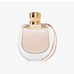 Un flacon de parfum en verre transparent avec un bouchon doré et un bracelet en cuir beige, étiqueté "Nomade Eau de Parfum" sur un fond blanc. Dakar