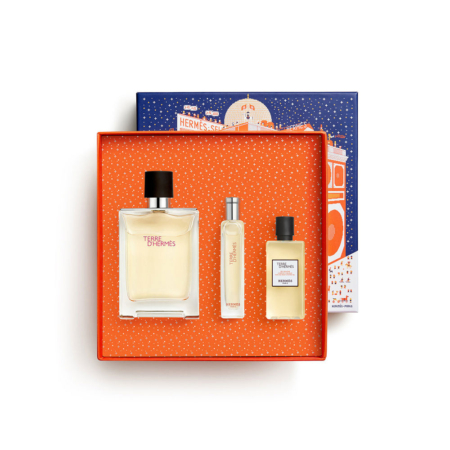 Un coffret cadeau Hermès orange à pois blancs contient trois parfums Terre d'Hermès : un grand flacon, un vaporisateur de voyage étroit et un petit flacon. Le couvercle de la boîte, sur lequel figure un bâtiment Hermès illustré, est placé derrière la boîte ouverte. Dakar
