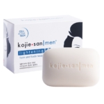 Une barre de savon éclaircissant blanc Kojie San Men est placée devant son emballage, qui présente un design minimaliste et une illustration partielle du visage d'un homme. Le savon est destiné à éclaircir la peau et à atténuer les taches brunes. Dakar