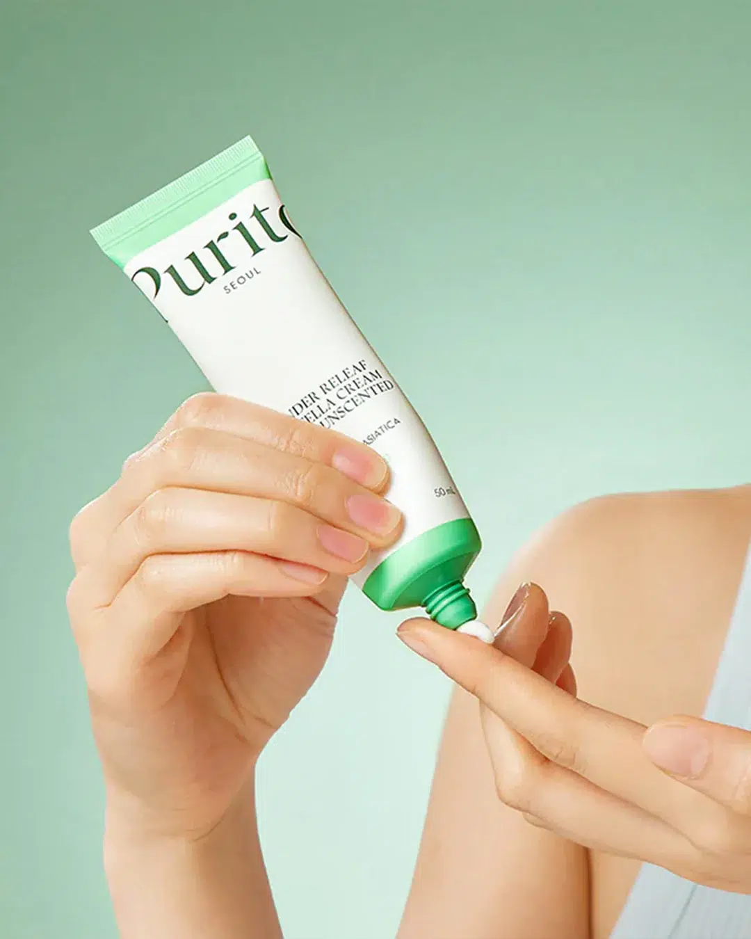 Une personne tient un tube de crème de soin Purito, en pressant une petite quantité sur le bout de son doigt, sur un fond vert clair. Seules les mains et une partie de l'épaule sont visibles. Dakar