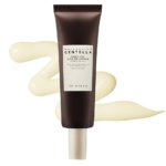 Un tube brun de SKIN1004 Madagascar Centella Glow Skin Sunblock SPF 50+ PA++++ est représenté à la verticale avec un tourbillon de produit crémeux jaune clair en arrière-plan. Dakar