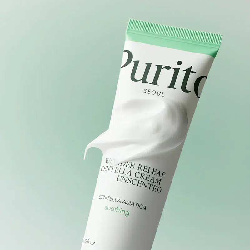 Un tube de crème Purito Wonder Releaf Centella Cream Unscented avec de la crème pressée hors de l'ouverture, sur un fond vert clair. Le tube est blanc et vert menthe avec un texte noir et vert. Dakar