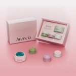 Un coffret de soins pour la peau de la marque Arencia est présenté sur une surface rose. La boîte ouverte présente trois pots de couleur verte, violette et blanche, avec des crèmes assorties sur le devant. L'emballage est minimal et élégant. Dakar