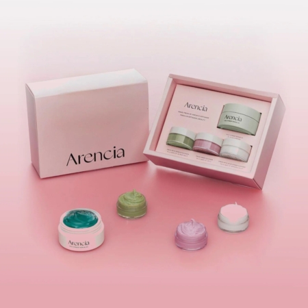 Un coffret de soins pour la peau de la marque Arencia est présenté sur une surface rose. La boîte ouverte présente trois pots de couleur verte, violette et blanche, avec des crèmes assorties sur le devant. L'emballage est minimal et élégant. Dakar