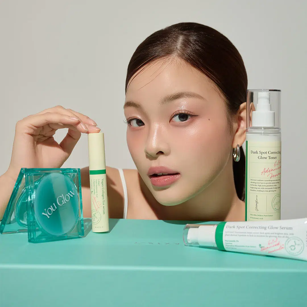 Une jeune femme à la peau éclatante pose avec plusieurs produits de soin de la peau dans un emballage vert clair et blanc, disposés sur une surface et une boîte turquoise. Les produits comprennent un sérum, un tonique, un stick et un compact étiqueté "You Glow". Dakar