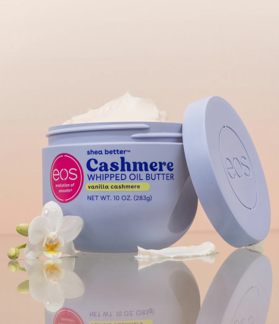 Un récipient lavande d'eos Cashmere Whipped Oil Butter est ouvert, montrant le produit crémeux à l'intérieur. Des fleurs d'orchidées blanches et une noisette de beurre se trouvent à proximité, sur un fond pastel doux. Dakar