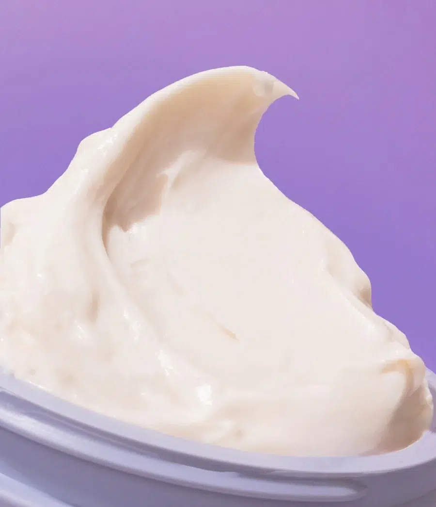 Gros plan d'un produit de soin crémeux, blanc, à la texture lisse et fouettée, sur un fond violet. Le produit semble être dans un récipient ouvert. Dakar
