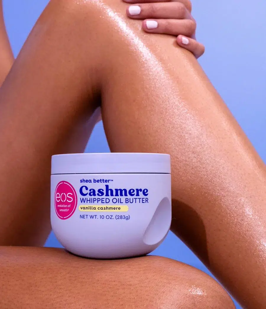 Les jambes lisses et hydratées d'une femme avec du vernis à ongles rose pâle sont montrées derrière un pot de beurre d'huile fouettée eos Cashmere, senteur cachemire vanille, sur un fond bleu doux. Dakar