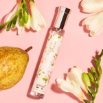 Un flacon de parfum floral étiqueté "Fleur de Poirier" par Adopt' repose sur un fond rose, entouré d'une poire fraîche et de fleurs de freesia blanches. Dakar