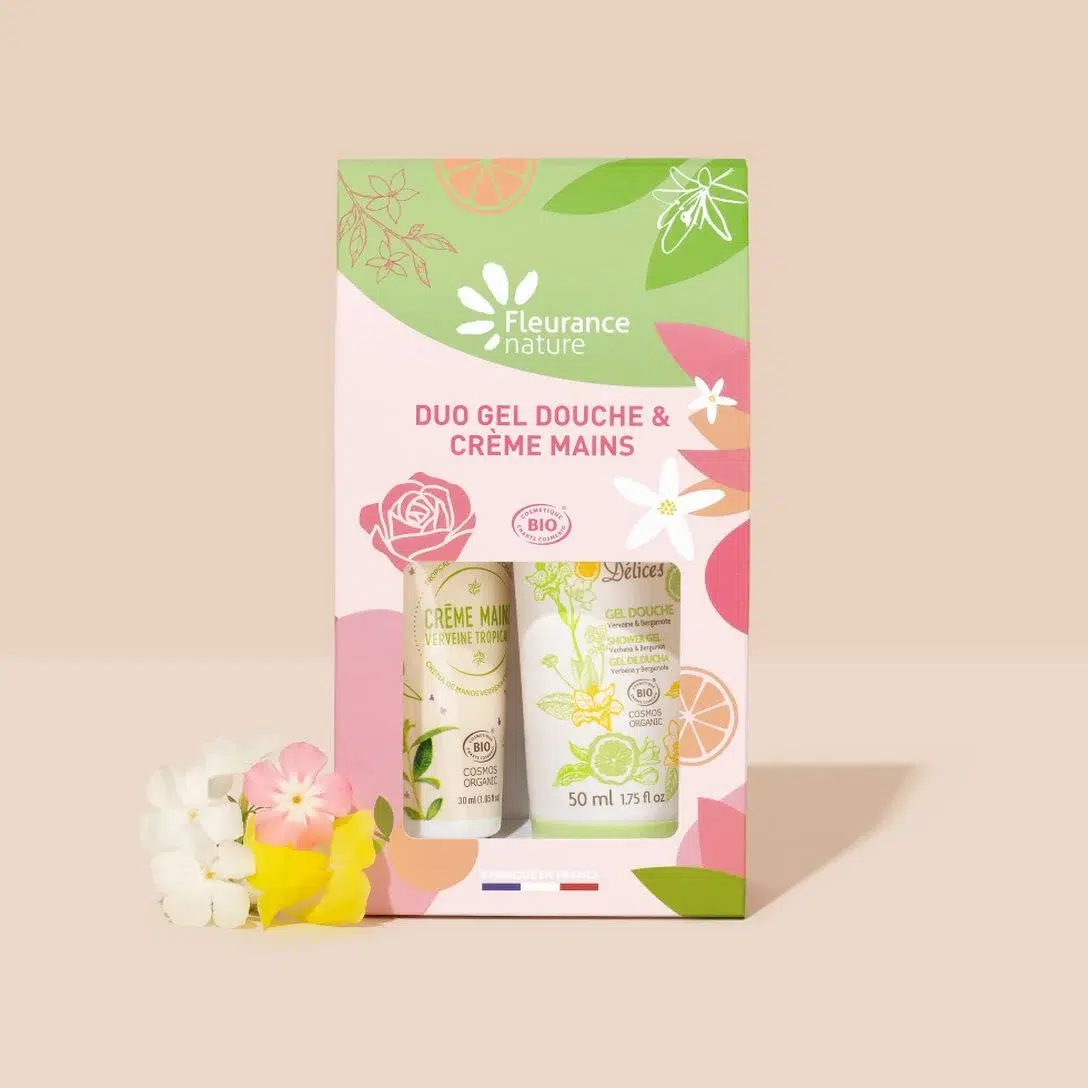 Coffret de produits Fleurance Nature, comprenant un duo de gel douche et de crème pour les mains. L'emballage est rose, vert et jaune avec des motifs floraux et d'agrumes, et une petite grappe de fleurs artificielles est placée à côté du coffret. Dakar