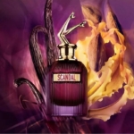 Un flacon de parfum étiqueté "SCANDAL" avec un bouchon doré en forme de jambes, sur un fond violet avec des gousses de vanille et des pétales de fleurs jaunes. Dakar