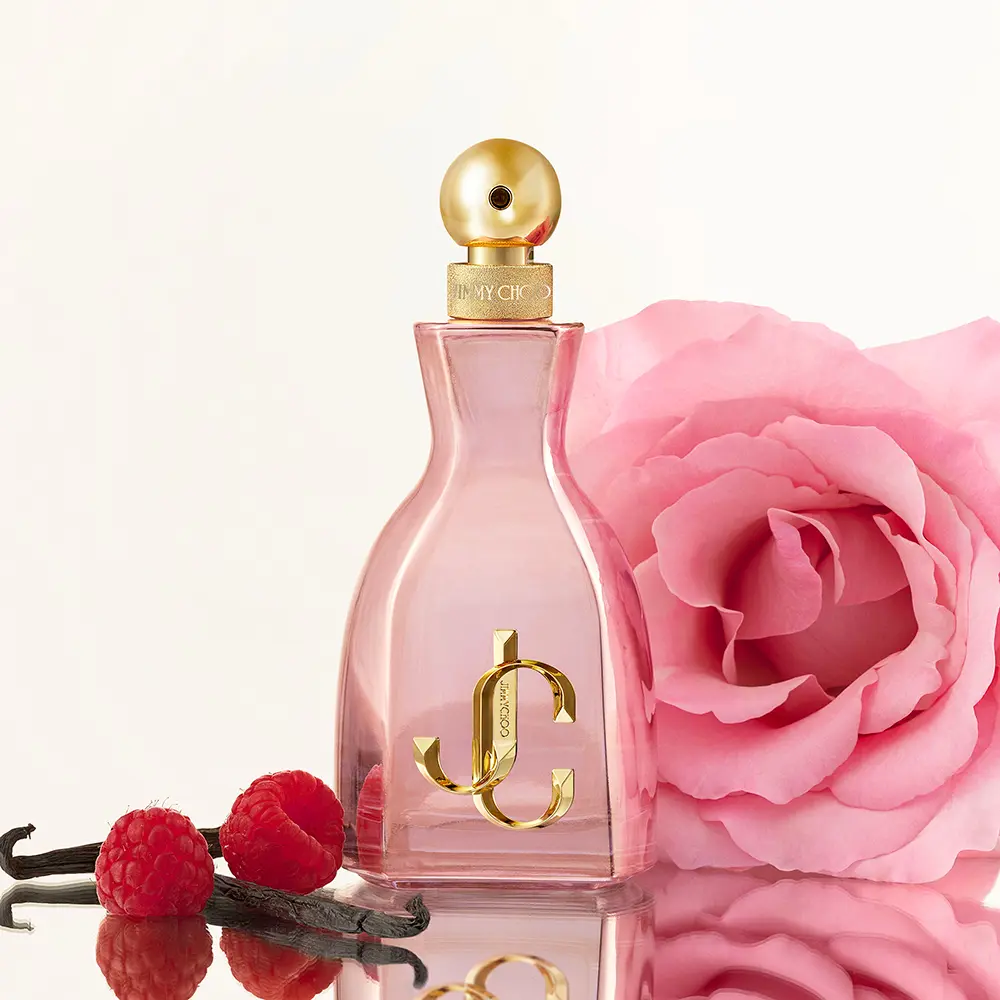 Un flacon de parfum rose avec un bouchon et un logo dorés se trouve à côté d'une rose douce, de deux framboises et d'une gousse de vanille sur une surface légère et réfléchissante. Dakar