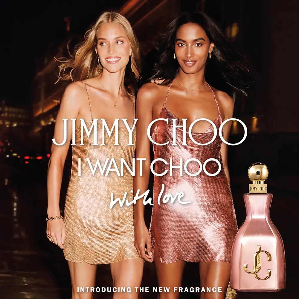Deux femmes vêtues de robes dorées et roses brillantes marchent ensemble en souriant dans la nuit. Le texte se lit comme suit : "Jimmy Choo I Want Choo With Love. Présentation du nouveau parfum". Un flacon de parfum rose est montré dans le coin. Dakar