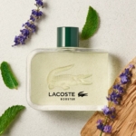 Un flacon de parfum Lacoste Booster avec un bouchon vert, entouré de brins de lavande, de feuilles vertes et d'un morceau de bois, présenté sur une surface légèrement texturée. Dakar