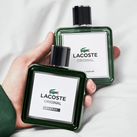 Une main tient deux flacons verts carrés de Lacoste Original Eau de Parfum vaporisateur avec des étiquettes blanches et des bouchons noirs, arborant le logo Lacoste. Une autre main tenant un deuxième flacon est visible à l'arrière-plan. Dakar