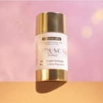 Un déodorant en stick solide, dont l'emballage or et blanc porte l'inscription "Musc Intime", est présenté sur un fond rose et or avec des effets de lumière douce et brillante. Dakar