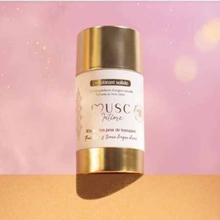 Un déodorant en stick solide, dont l'emballage or et blanc porte l'inscription "Musc Intime", est présenté sur un fond rose et or avec des effets de lumière douce et brillante. Dakar