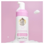 Un flacon rose et blanc de Musc Intime Foaming Cleanser repose sur une mousse blanche, avec plus de mousse sur la pompe et flottant à côté. Le flacon comporte un texte en or et en rose ainsi qu'un label de prix de beauté. Dakar