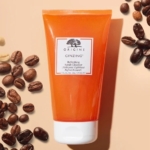 Un tube orange de Origins Ginzing Nettoyant 2 en 1 Exfoliant Rafraîchissant 150 ml se tient debout sur une surface beige, avec des grains de café entiers et quelques grains décortiqués éparpillés autour de lui. Dakar