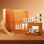Une collection de produits de soin de la peau, comprenant des flacons, des tubes et des pots, est soigneusement disposée devant une boîte de calendrier de l'avent ouverte, orange et dorée, avec des compartiments numérotés, le tout sur un fond brun chaud. Dakar
