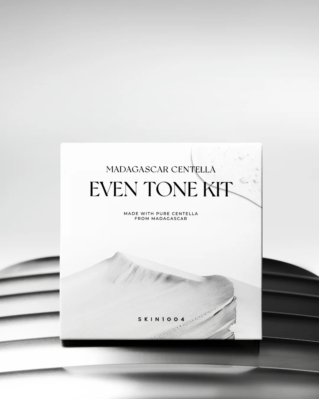 Une boîte blanche étiquetée "Skin1004 Kit Unifiant le Teint à la Centella de Madagascar" repose sur une surface métallique incurvée, présentant des graphiques minimalistes et un design propre et moderne. Dakar