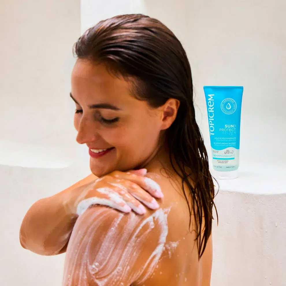 Une femme aux cheveux mouillés sourit en appliquant de la crème solaire sur son épaule. Un tube de crème solaire Topicrem SUN Protect est visible en arrière-plan sur une surface blanche. Dakar