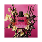 Un flacon de parfum Valentino rose au design texturé et clouté est entouré de fleurs jaunes et de baies sombres sur un fond rose vif. Dakar