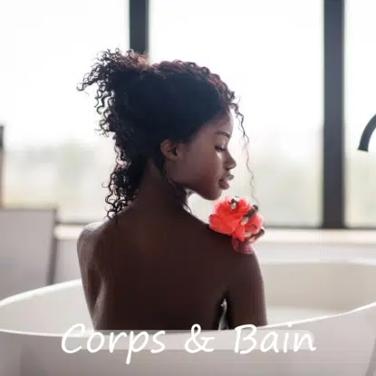Une femme aux cheveux bouclés est assise dans une baignoire blanche, se lavant doucement l'épaule avec une éponge de bain rouge. Une douce lumière naturelle traverse les grandes fenêtres derrière elle, créant un moment de sérénité Univers Cosmetix. La mention "Corps & Bain" est inscrite au bas de l'image. Dakar